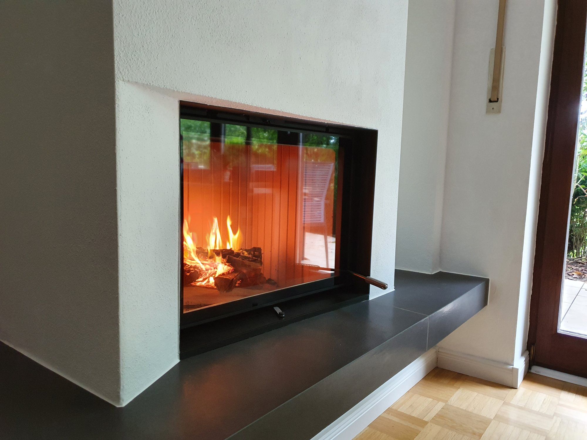 Brennendes Holzfeuer hinter der Sichtscheibe eines weiß verputzten Heizkamins mit dunkler Steinbank