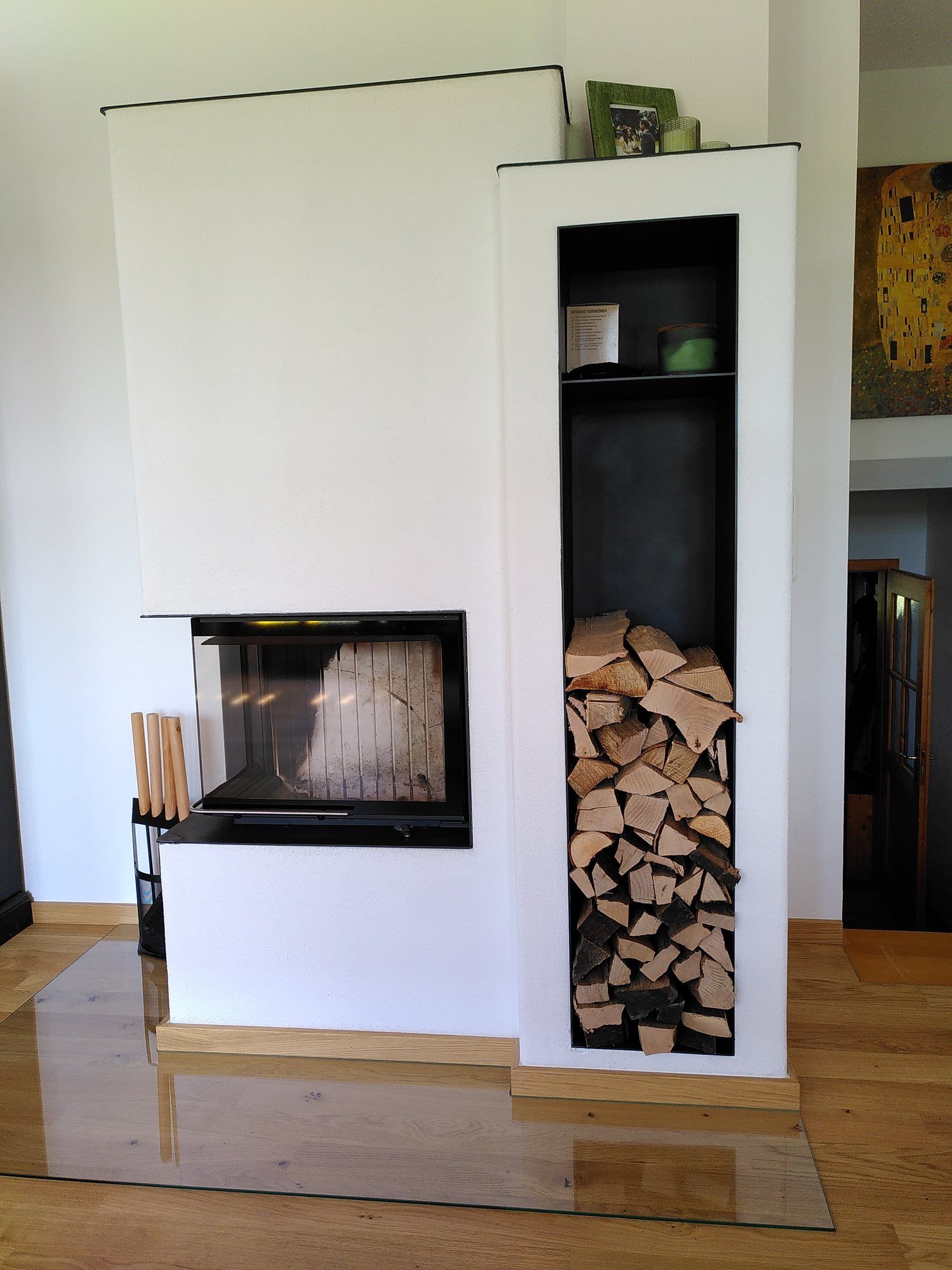 Weißer Heizkamin mit Eck-Sichtscheibe und hohem, mit Brennholz gefülltem Holzfach
