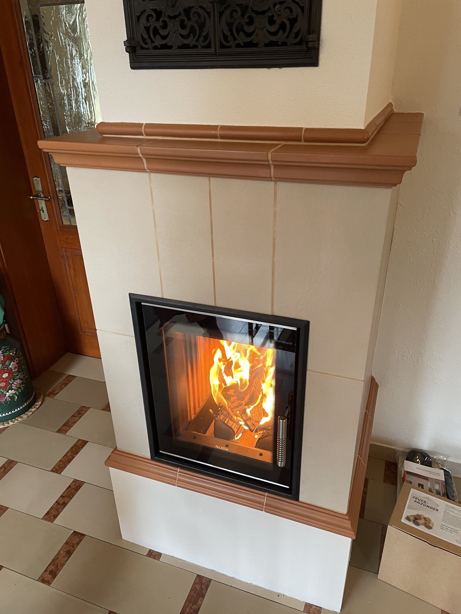 Nahaufnahme eines Kachelofens: cremeweiße Kacheln, profiliertes Terrakotta-Gesims und Feuer im Brennraum